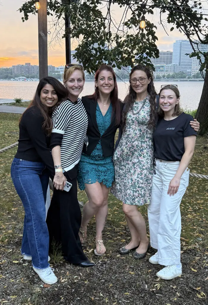 Five women smiling and posing together outside by a river with city buildings in the background at sunset.