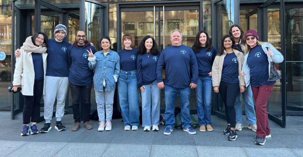 A group of people in matching blue shirts pose together and smile outside a glass building entrance.