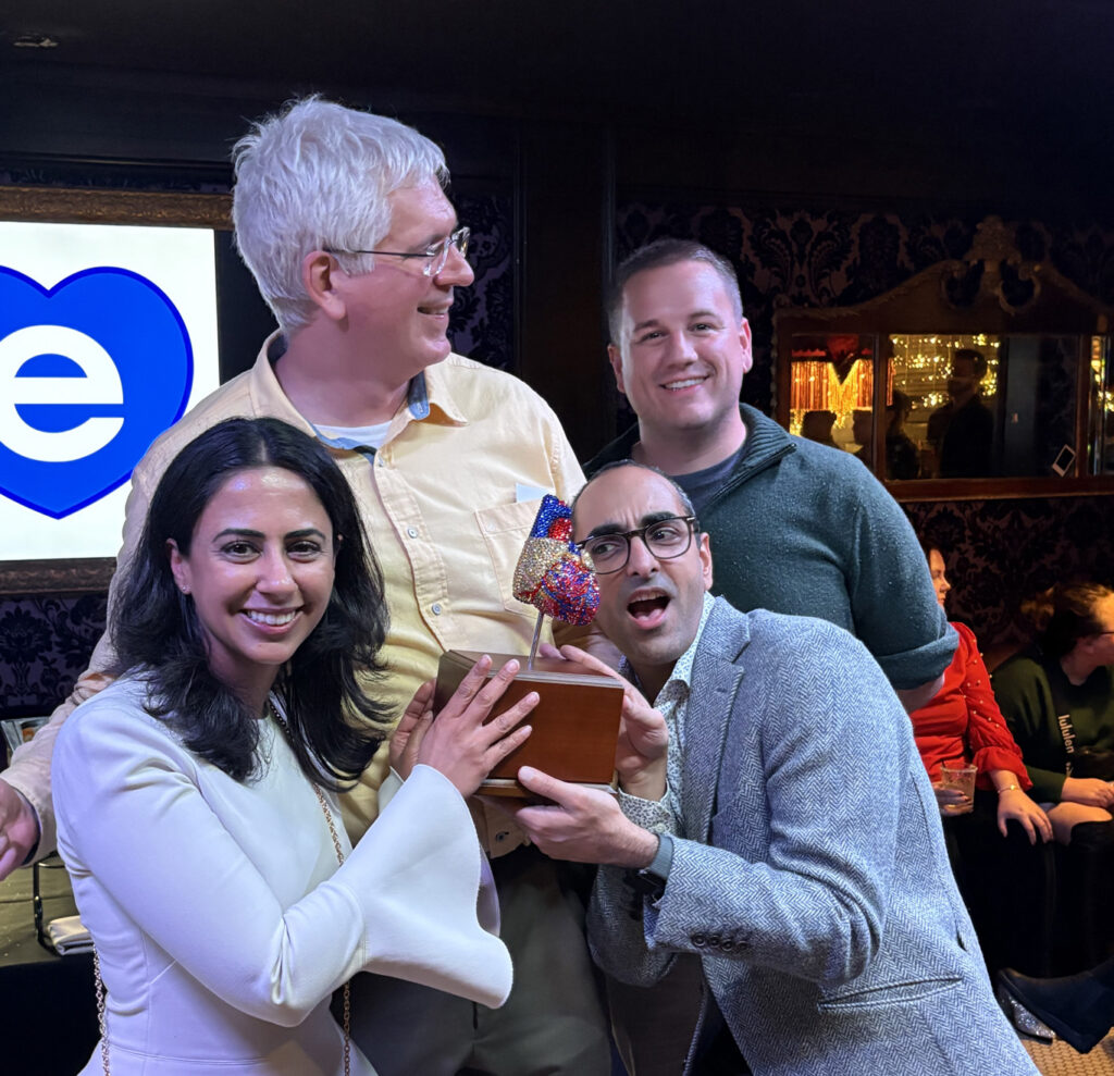Four people smiling and holding a heart-shaped trophy at an indoor event, with others in the background.