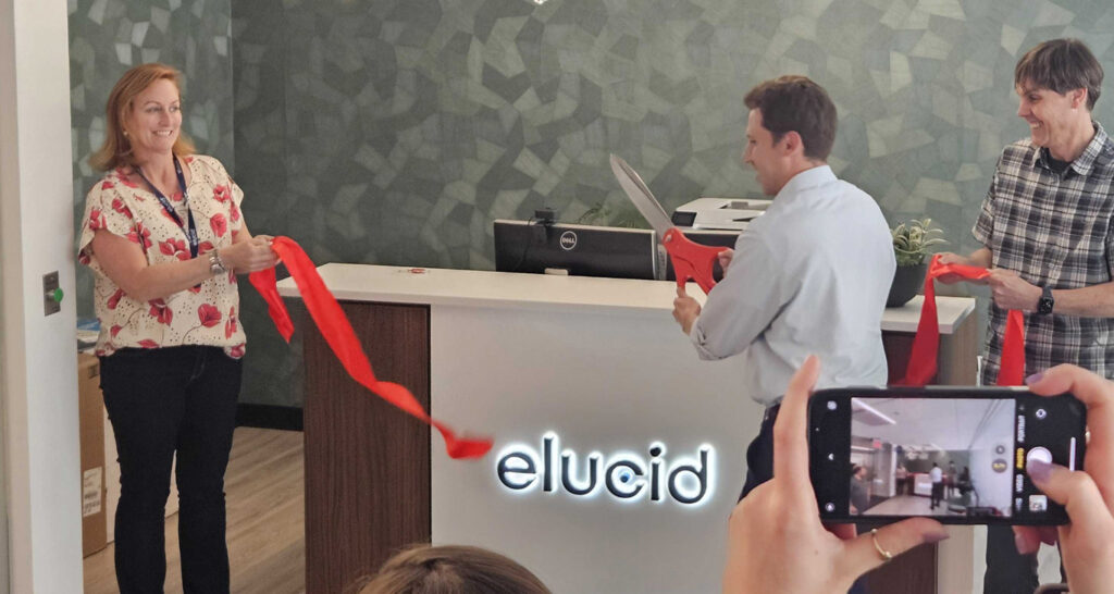 Three people at an Elucid office hold and cut a red ribbon; someone photographs the scene with a phone.