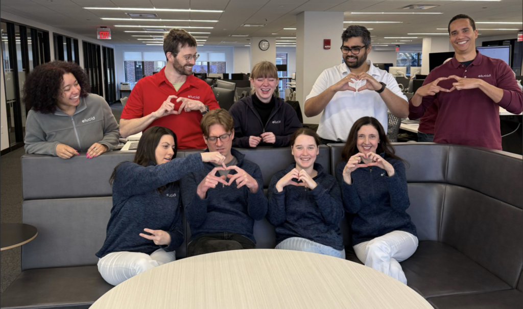 Eight people in an office sit and stand together, smiling and making heart shapes with their hands.