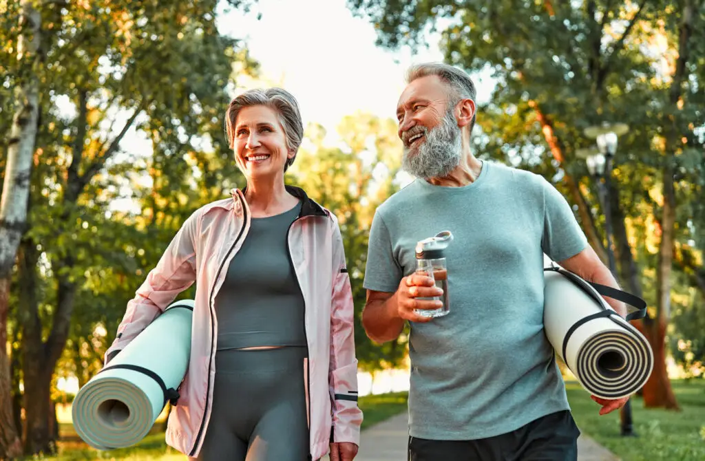Smiling older couple carrying yoga mats and a water bottle, walking together in a sunny park.