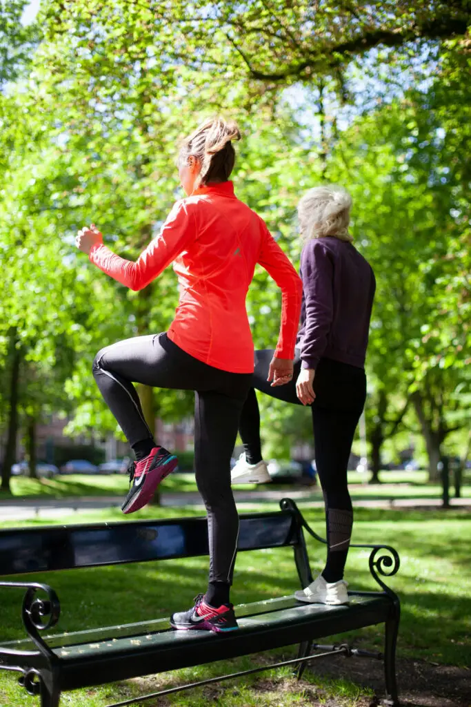 Two women exercising on a park bench, one stepping up while the other stands beside her, surrounded by greenery.